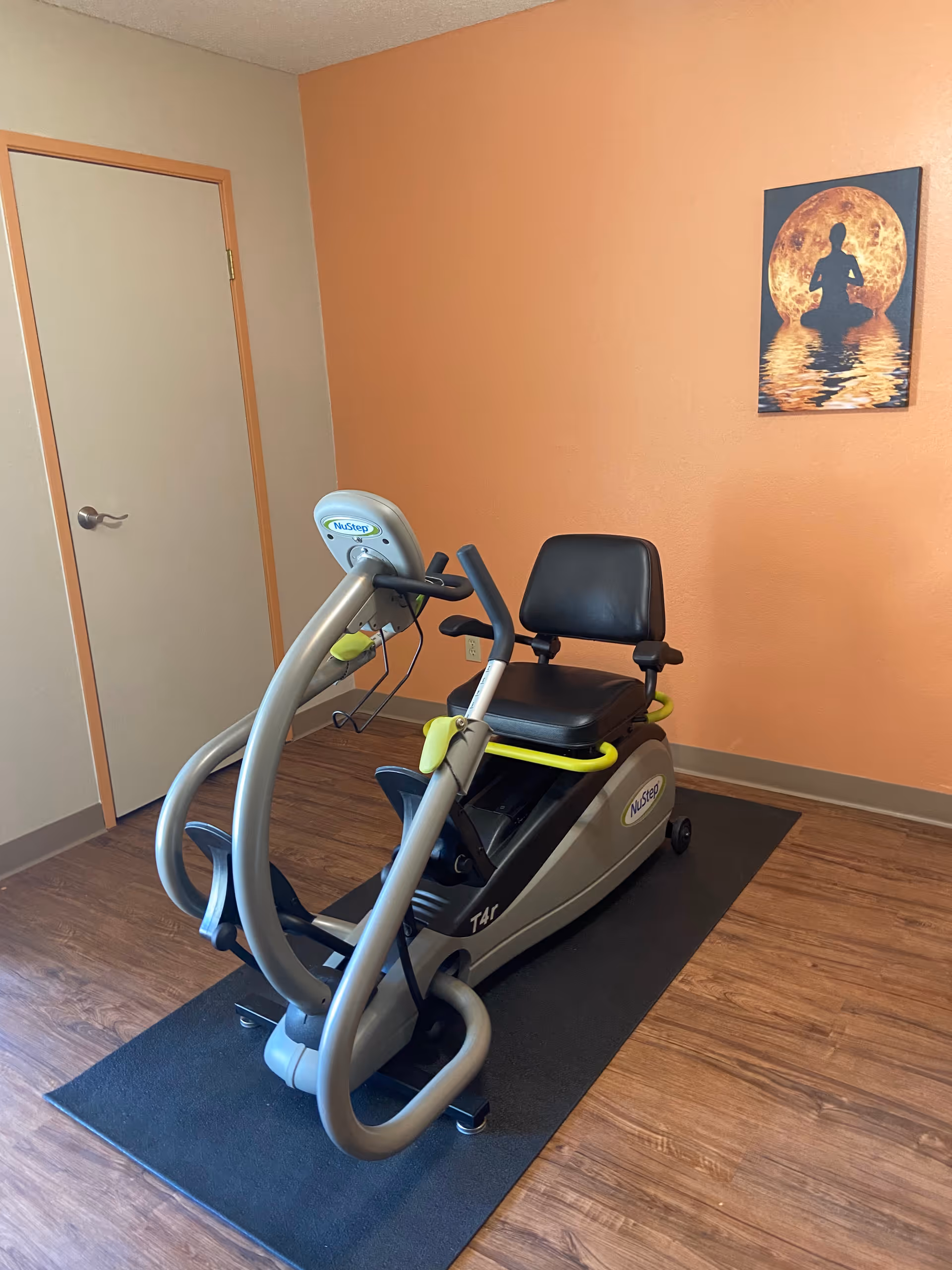 A NuStep exercise machine with a black seat and handles is placed on a black mat in a room with wood flooring. The walls are painted beige and orange, and there is a door on the left side. A framed picture of a person meditating in front of a large orange moon hangs on the orange wall.