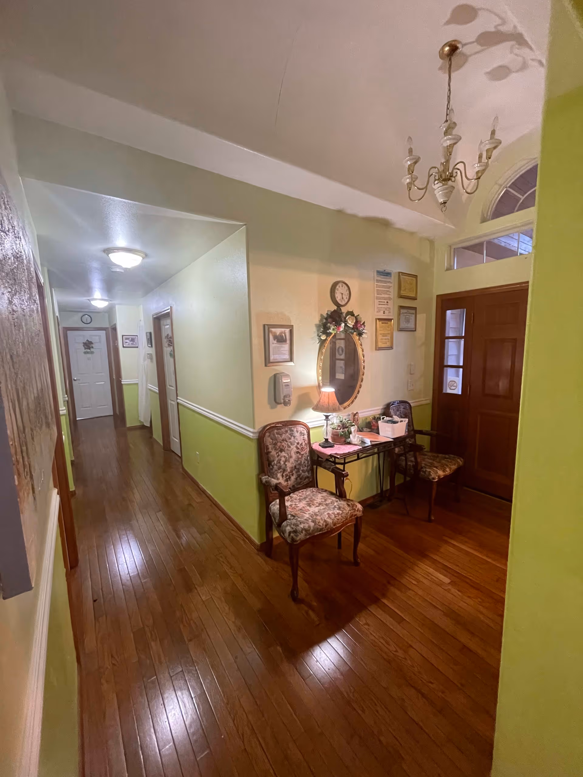 Interior hallway of a senior living facility with wooden flooring, light green and cream walls, two floral upholstered chairs, a small table with a lamp and plants, a decorative oval mirror with a floral wreath, a clock, framed certificates on the wall, and a wooden front door with glass panels. A chandelier hangs from the ceiling.