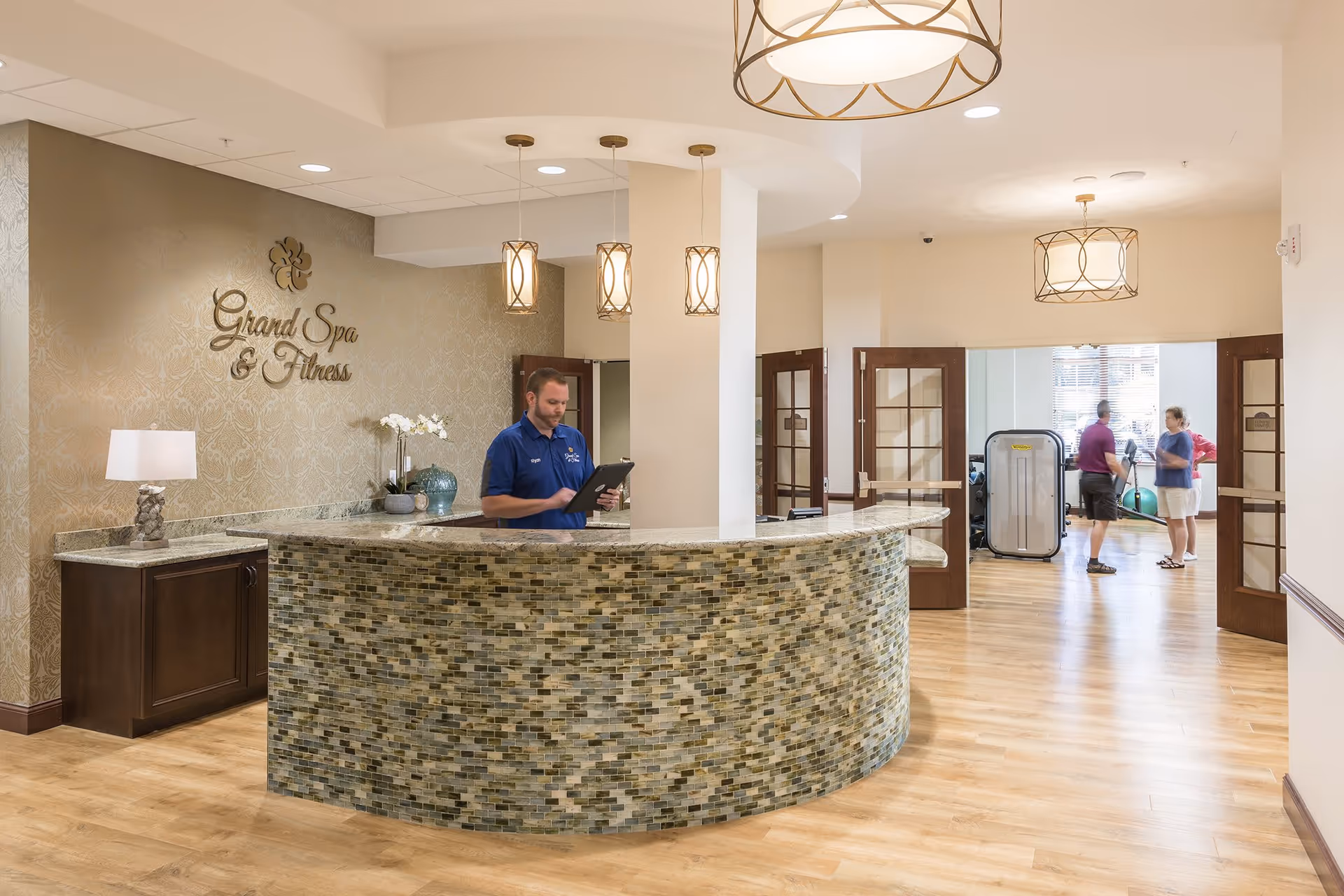 Reception area of Grand Spa & Fitness with a curved mosaic tile front desk, a staff member in a blue shirt using a tablet, pendant lights hanging from the ceiling, and two people conversing in the fitness room visible through open double doors.