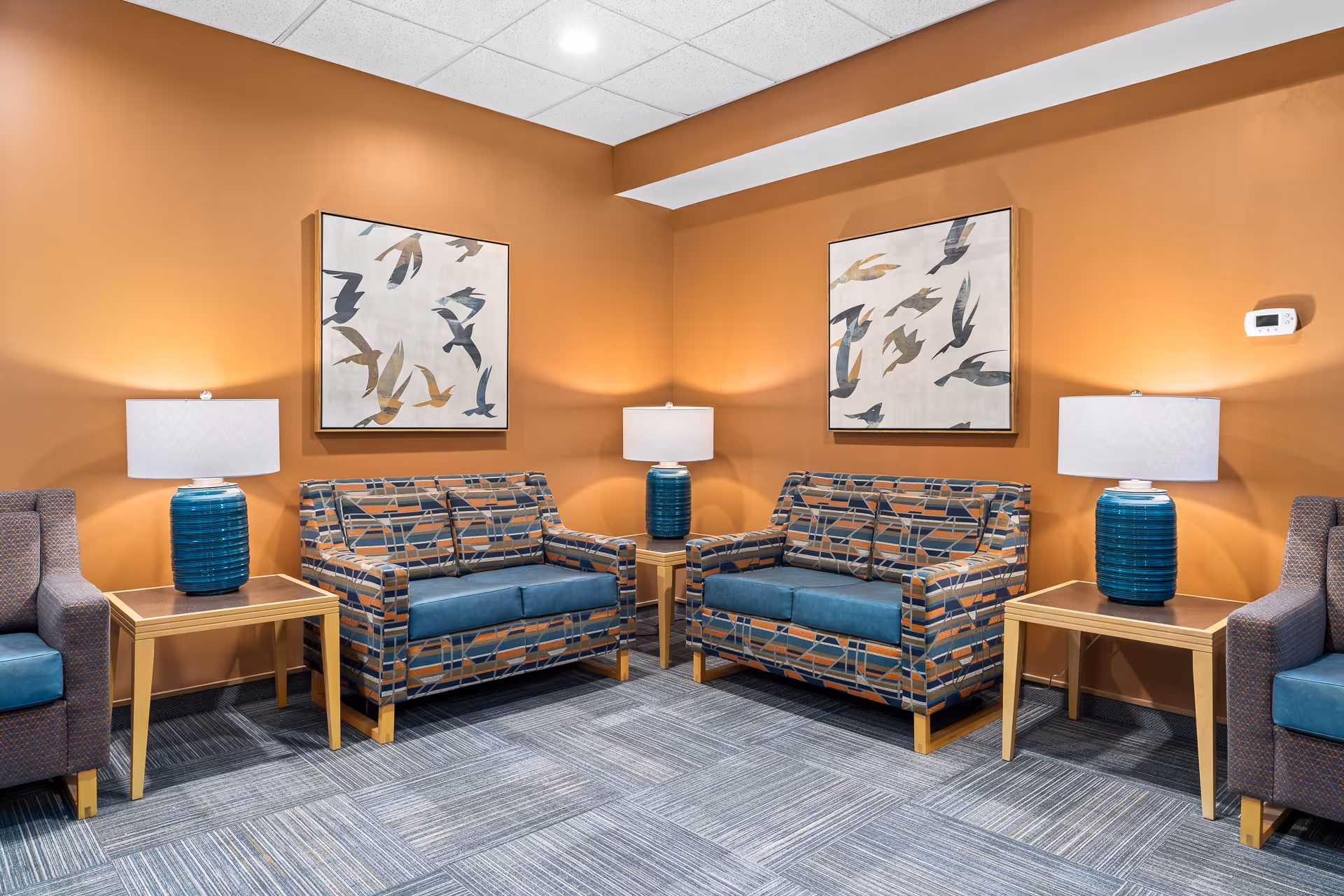 A cozy seating area with two patterned loveseats and two armchairs arranged around two wooden side tables with blue ceramic lamps. The walls are painted a warm orange color and decorated with two framed artworks featuring abstract bird designs. The floor is carpeted with a gray patterned carpet, and the ceiling has recessed lighting.
