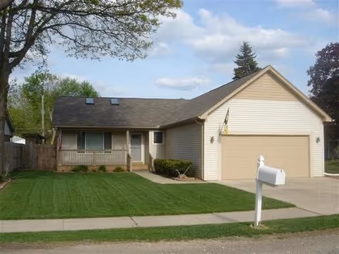 Single-story house with beige siding, a two-car garage, a small front porch, and a well-maintained green lawn with a white mailbox near the sidewalk. Trees and a partly cloudy sky are visible in the background.