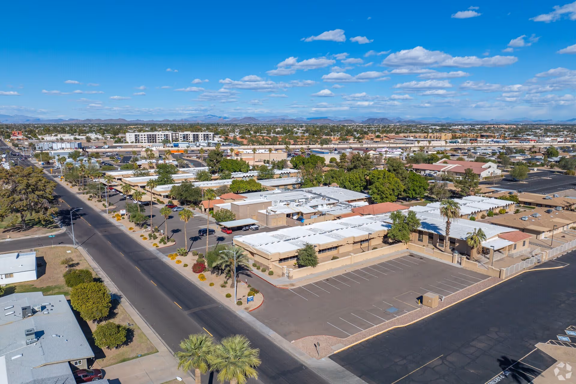 Aerial view of LifeStream at Sun City facility showing multiple single-story buildings with white roofs, surrounded by parking lots, trees, and nearby streets under a blue sky with scattered clouds.