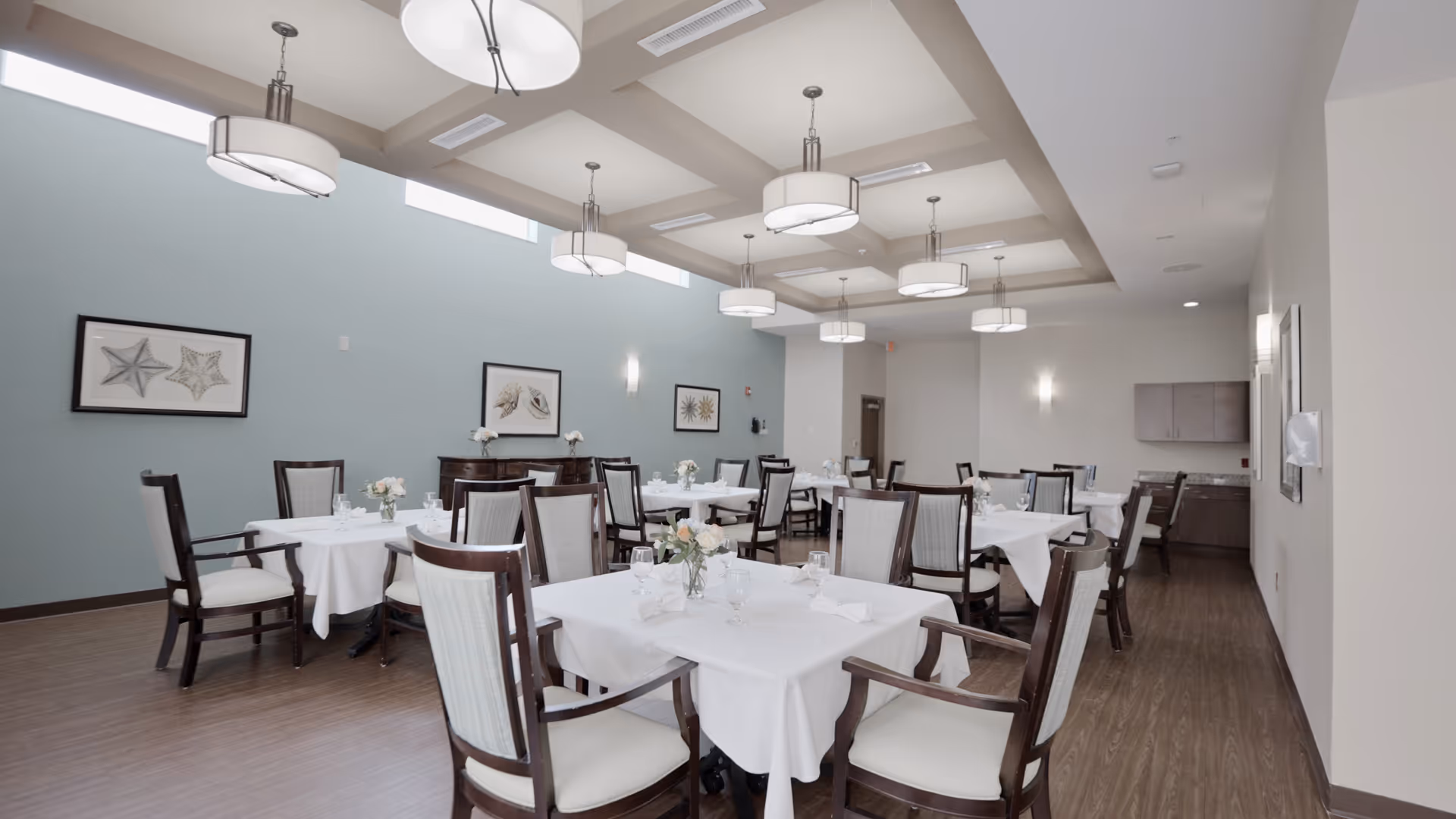 A bright dining room with several tables covered in white tablecloths, each set with glassware, napkins, and small floral centerpieces. The room has wooden chairs with cushioned seats and backs, light blue walls adorned with framed artwork, and modern ceiling lights hanging from a coffered ceiling. The floor is wood, and there is a sideboard and cabinetry along the walls.
