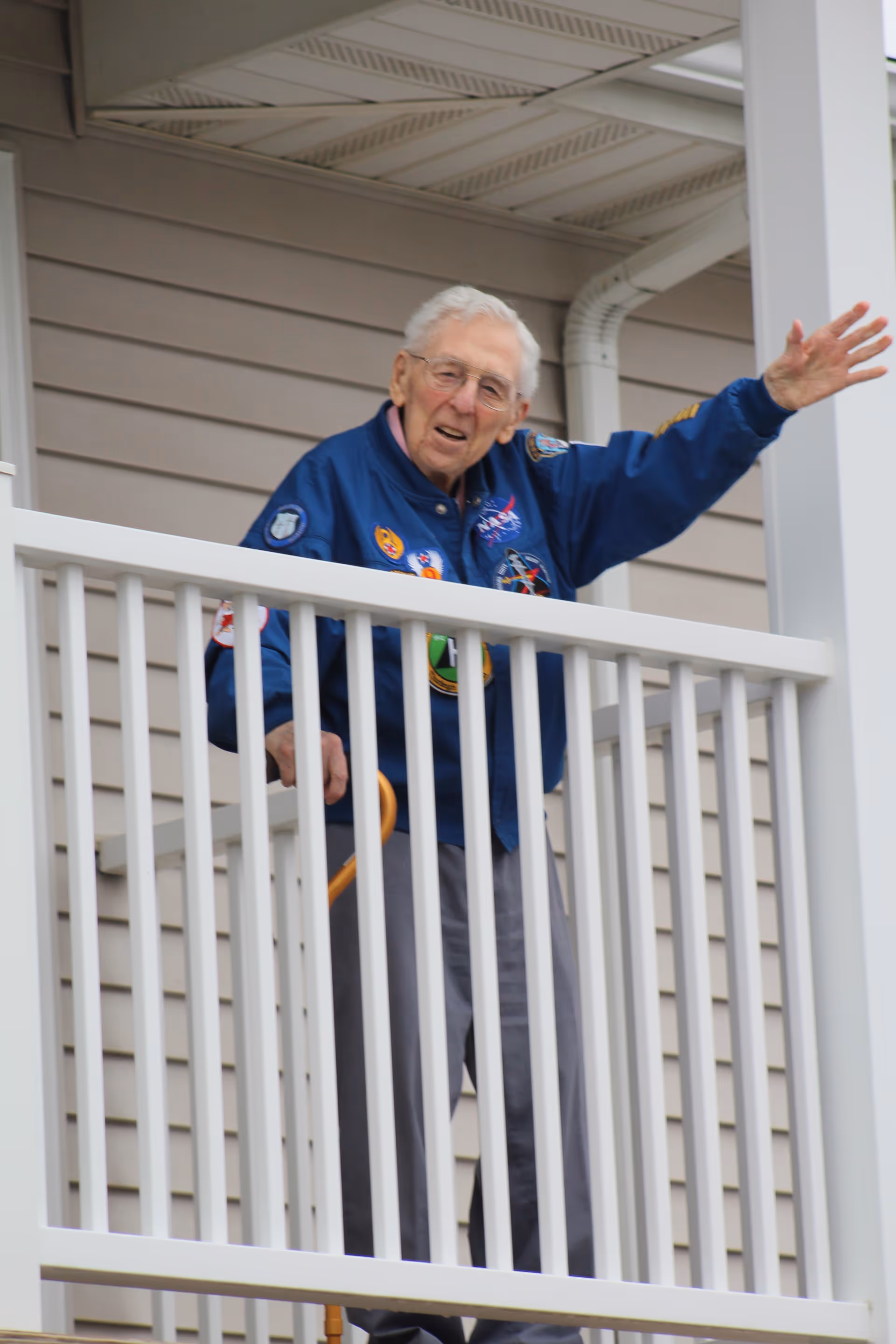 An elderly man wearing a blue jacket with various patches stands on a balcony with white railings, waving with his right hand. The building exterior has beige siding and white trim.
