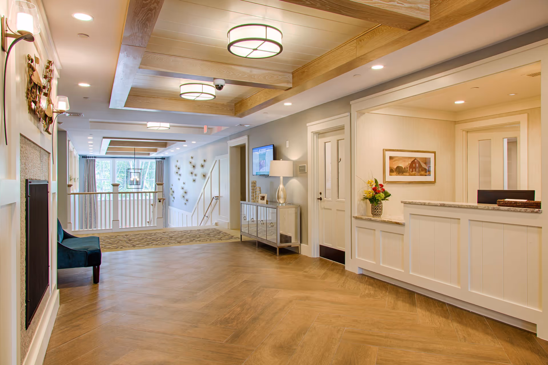 A bright and spacious interior hallway of a senior living facility with wood flooring, a reception desk with a flower arrangement, a wall-mounted TV, decorative wall art, and a seating area near a fireplace. The ceiling features wooden beams and modern light fixtures.