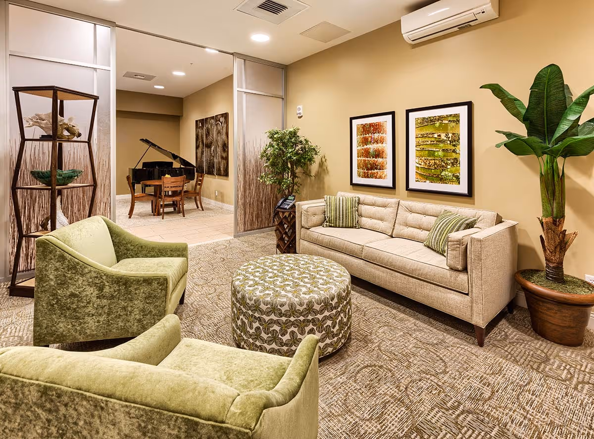 A cozy living room area with a beige sofa, two green armchairs, and a round patterned ottoman. The walls are painted light beige and decorated with two framed artworks. There is a tall potted plant in the corner and a wooden shelving unit with decorative items. In the background, there is a small dining area with a table, chairs, and a grand piano.