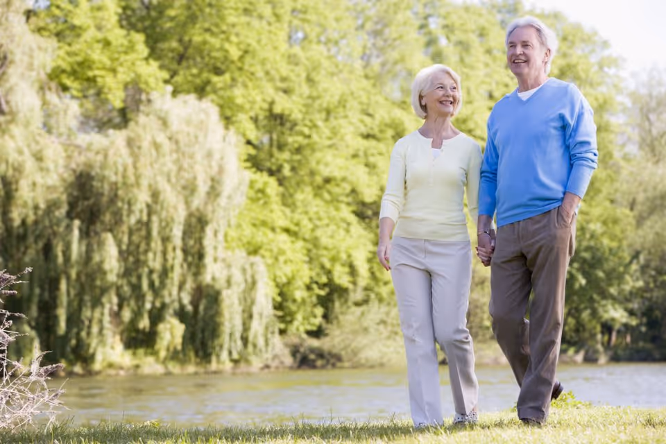 An elderly couple holding hands and walking outdoors near a body of water with lush green trees in the background on a sunny day.