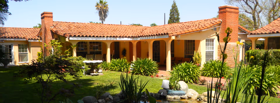 Exterior view of a single-story senior living facility with a red tiled roof, beige walls, and two brick chimneys. The building is surrounded by a well-maintained garden with green shrubs, plants, a small pond with rocks, and a birdbath. Tall trees and a clear blue sky are visible in the background.