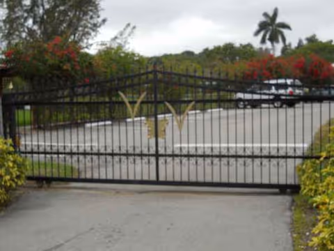 A wrought iron gate opens to a driveway with landscaped shrubs.