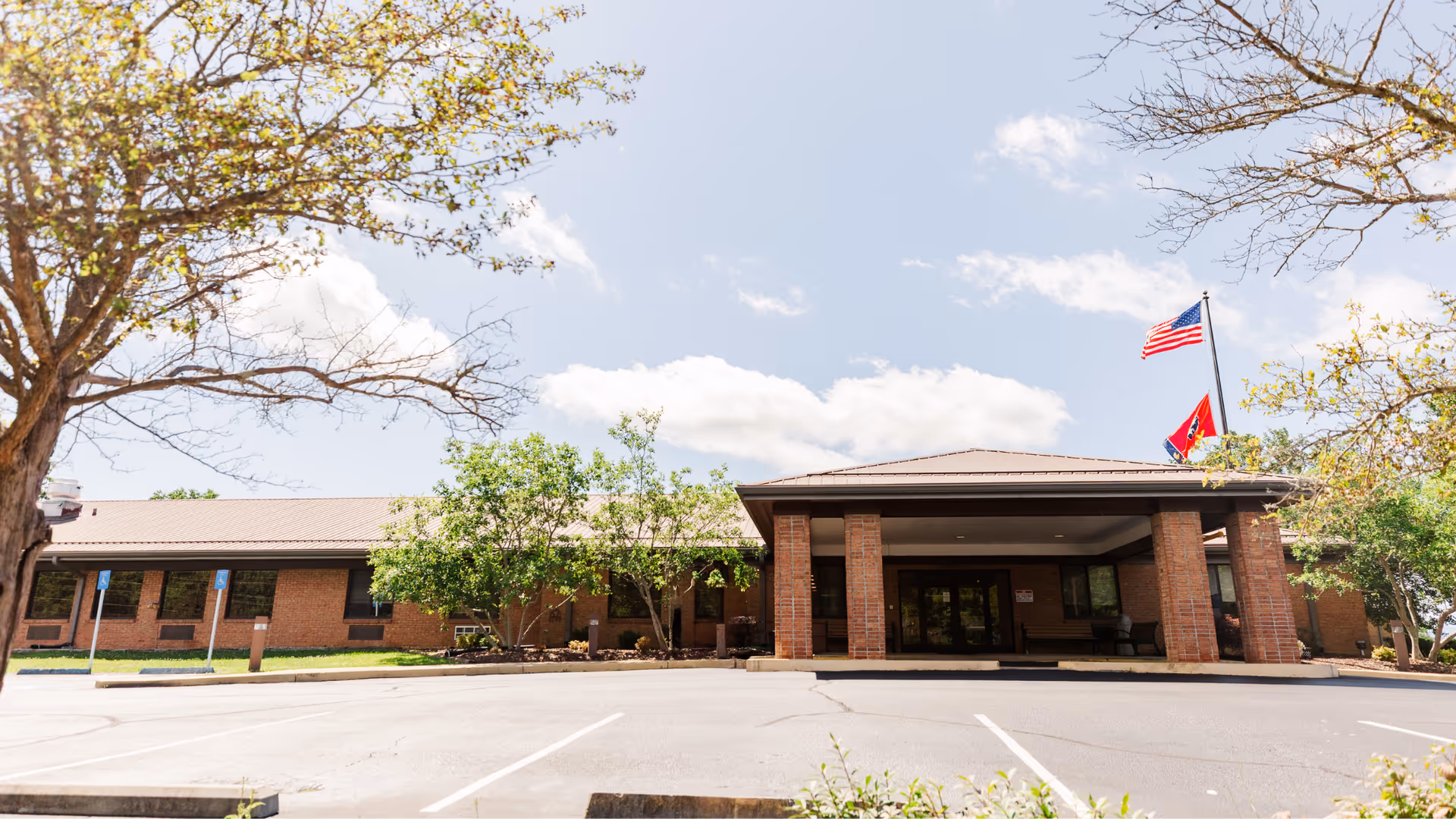 Front exterior of a single-story brick healthcare building with a covered entrance, flagpoles, trees, and an empty parking lot.