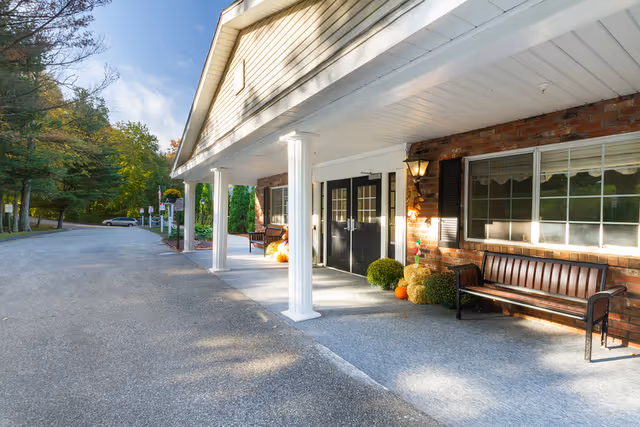 Covered entrance of a red-brick building with white columns, benches, double doors, pumpkins, and a driveway lined with trees.