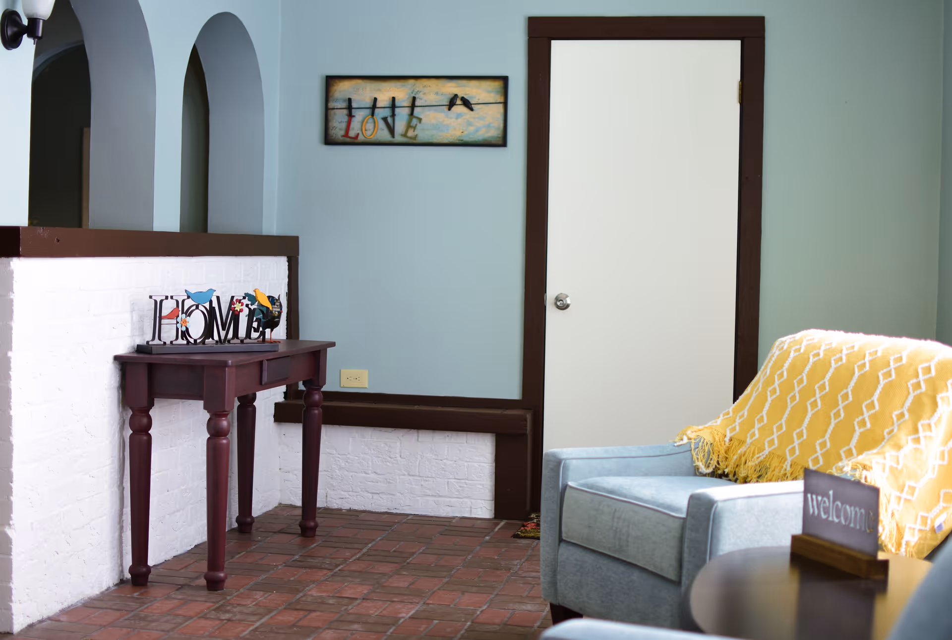 A cozy corner of a room with a blue armchair draped with a yellow patterned blanket, a small round table with a 'welcome' sign, a wooden console table with a decorative 'HOME' sign, light blue walls with dark brown trim, a white door, and a wall art piece with the word 'LOVE' and birds on a wire.