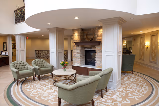 A cozy seating area in a senior living facility with four green upholstered chairs arranged around a round marble-top coffee table. The space features a stone fireplace with a wooden mantle and decorative horseshoe above it. The area is carpeted with a patterned rug and surrounded by white columns and soft lighting.