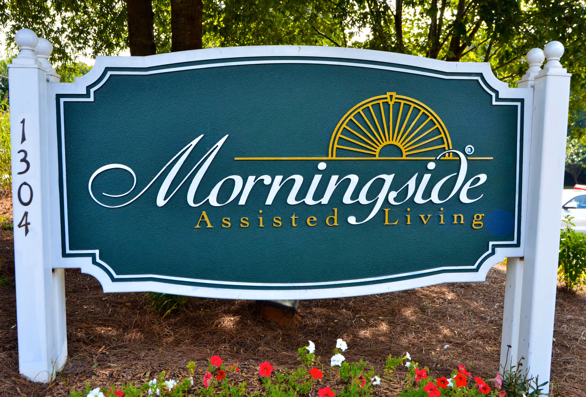 A green and white sign for Morningside Assisted Living with a yellow sun logo above the text, mounted on white posts with the number 1304 on the left post, surrounded by mulch and red and white flowers at the base, with trees and parked cars in the background.