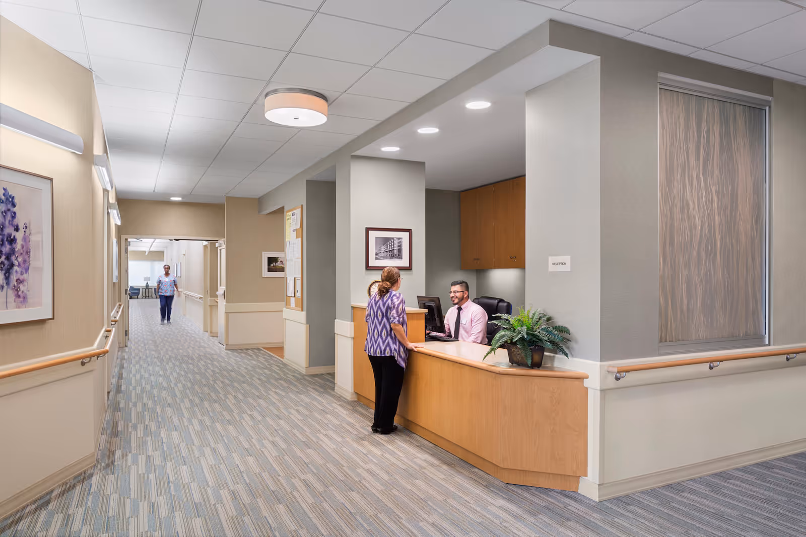 Interior view of a senior living facility hallway with a reception desk on the right. A woman is standing at the reception desk talking to a seated receptionist. The hallway is carpeted with a patterned design and has handrails along the walls. There is a person walking down the hallway in the background. The walls are decorated with framed artwork and bulletin boards.