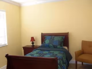 A small bedroom with a single wooden bed featuring a blue and green patterned bedspread and matching pillow. Next to the bed is a wooden nightstand with a red lamp. There is a brown upholstered chair to the right of the bed. The walls are painted light yellow, and there is a window with white blinds on the left side.