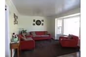 A living room with two red sofas and one red armchair arranged around a dark carpet. There is a small wooden side table with a lamp and decorative items on the left side. The walls are light-colored with a clock and framed pictures hanging above the sofa. A large window on the right side lets in natural light.