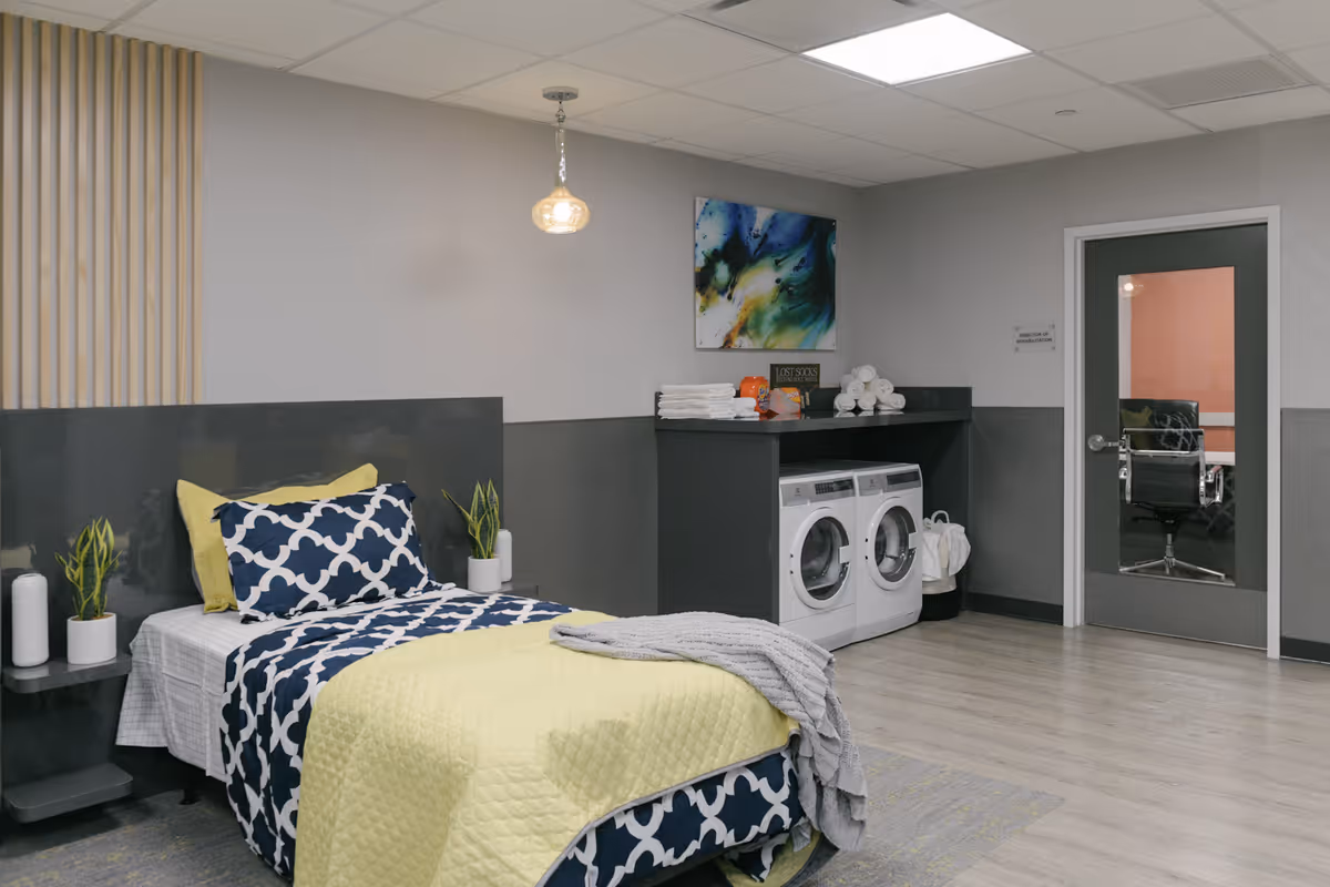 A room with a single bed made with patterned navy and white bedding and a yellow quilt. There are two small potted plants on a small table next to the bed. Across from the bed is a laundry area with a washer and dryer under a counter, which has folded towels and some decorative items on top. A modern abstract painting hangs on the wall above the laundry area. The room has light wood flooring and gray walls with a door leading to another room.
