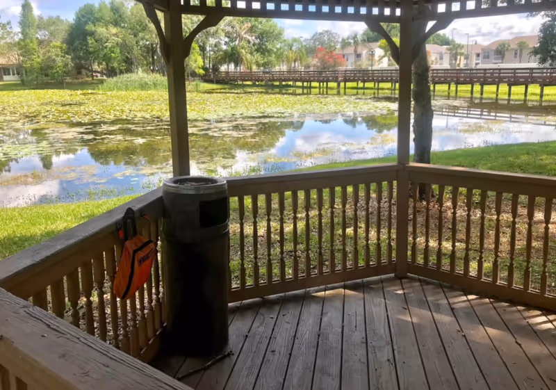 View from a wooden gazebo overlooking a pond covered with lily pads. There is a wooden railing around the gazebo and a trash can with an orange bag hanging on the side. In the background, there are trees, a wooden bridge, and residential buildings under a partly cloudy sky.