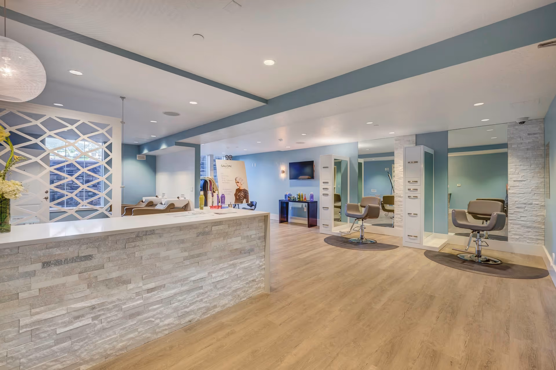 Interior view of a modern salon area in The Ivy at Draper facility featuring a reception counter with a stone facade, two styling chairs in front of large mirrors, shampoo stations in the background, and a display of hair care products. The room has light wood flooring, blue walls, and recessed ceiling lights.