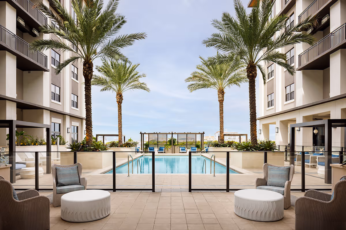 Sunlit outdoor pool courtyard framed by palm trees and lounge seating between two building wings.