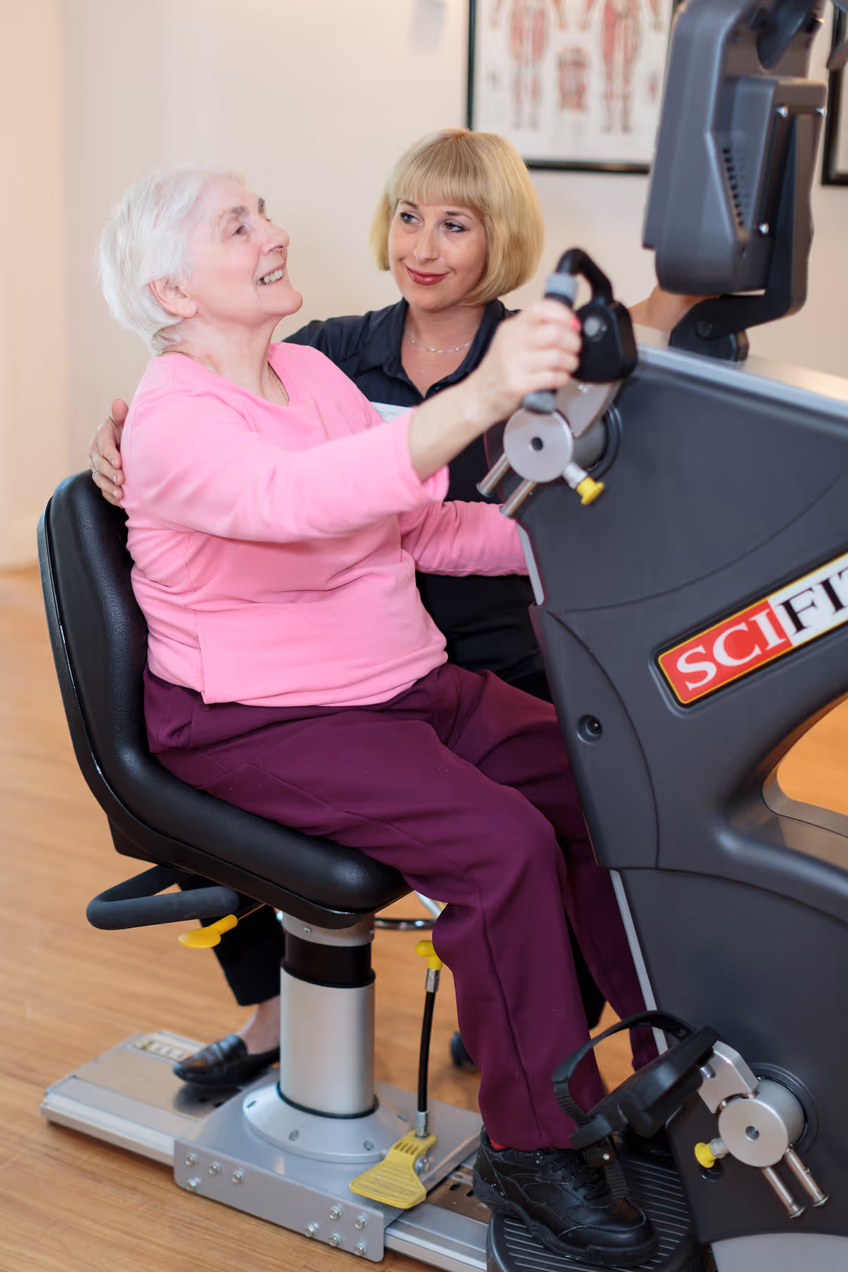 An elderly woman in a pink sweater and purple pants is seated on a recumbent exercise bike, smiling and holding the handles. A caregiver with short blonde hair is sitting beside her, offering support and encouragement. The setting appears to be indoors with a wooden floor and a medical anatomy poster on the wall in the background.