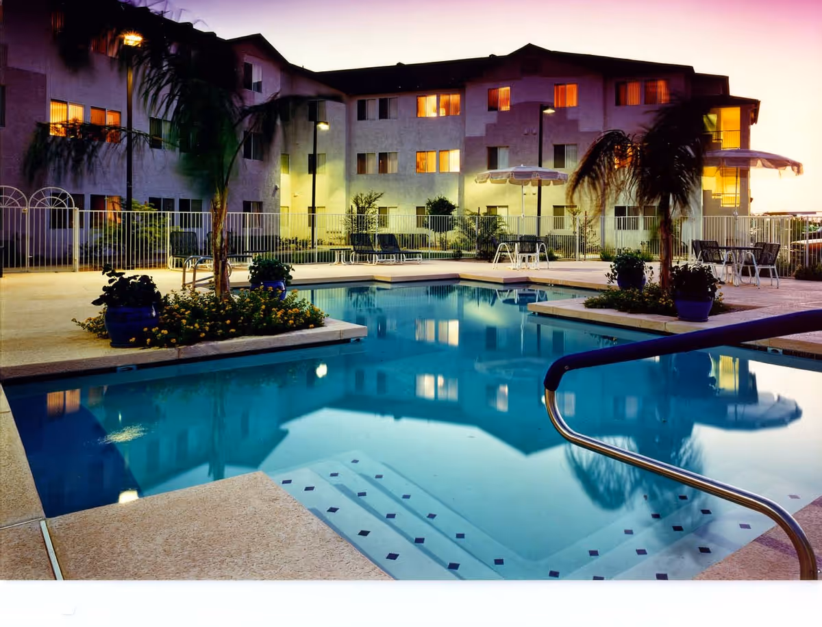Outdoor swimming pool area at dusk with a multi-story building in the background. The pool is surrounded by a concrete deck with potted plants, palm trees, lounge chairs, and tables with umbrellas. The building windows are lit, and the pool water reflects the building and surrounding lights.