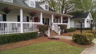 Exterior view of a white building with a covered porch, white railings, and steps leading up to the entrance. There are hanging flower baskets, trimmed bushes, a lamp post, and a well-maintained lawn in front.