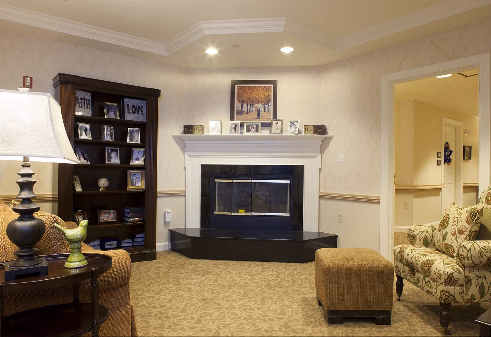A cozy living room area with a fireplace centered on the back wall. Above the fireplace is a framed picture and several small photo frames and decorative blocks with words like 'Live' and 'Love'. To the left is a dark wooden bookshelf filled with framed photos and decorative items. On the right side, there is a floral patterned armchair and a brown ottoman. A table lamp with a green bird figurine sits on a small round table next to a brown sofa. The walls are light-colored with a subtle pattern, and the carpet has a beige floral design. An open doorway leads to a hallway in the background.