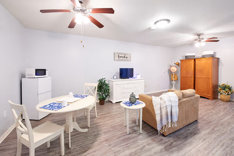 A bright and spacious living area with light gray walls and wood flooring. The room features a small beige couch with a white and blue throw blanket, a round side table with a decorative item, a white dining table with two chairs and blue placemats, a white cabinet with a small TV and clock, a potted plant, a wooden wardrobe, and a small refrigerator with a microwave on top. Ceiling fans with lights are mounted on the ceiling.