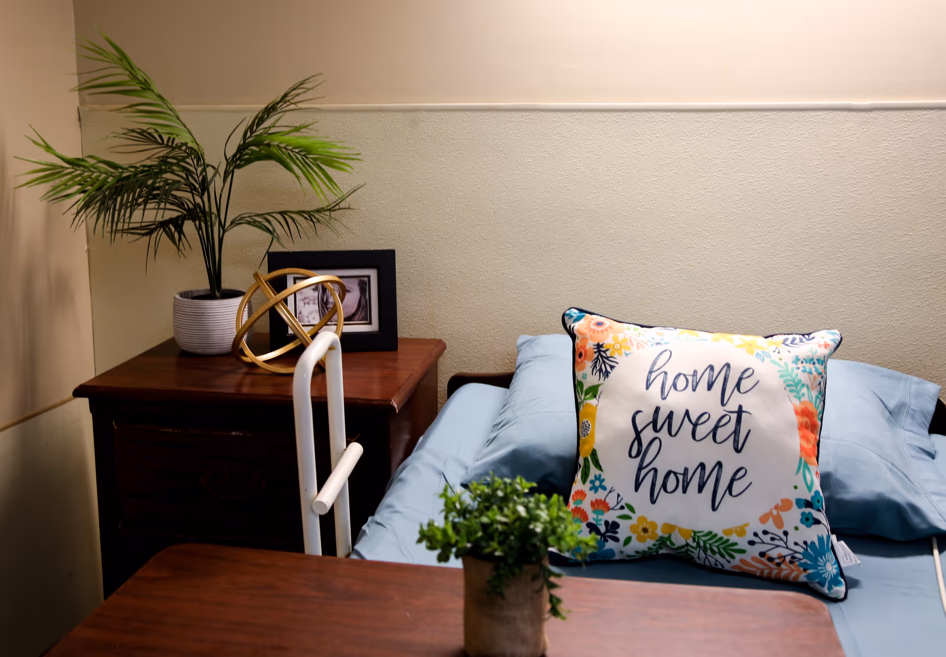 A cozy bedroom corner in a senior living facility featuring a bed with light blue bedding and a decorative pillow that says 'home sweet home'. Next to the bed is a wooden nightstand with a potted plant, a framed photo, and a decorative golden orb. A small green plant is also visible on a wooden table in the foreground.