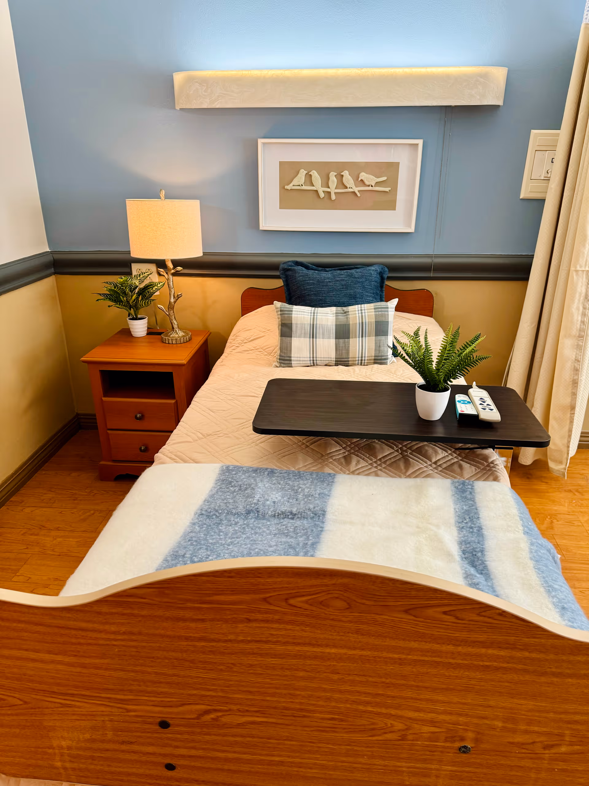 A neatly made single bed in a nursing facility room with a wooden headboard and footboard. The bed has a beige quilt, a blue and white plaid pillow, and a dark blue cushion. A small wooden bedside table with a lamp and a potted plant is next to the bed. Above the bed is a framed artwork of birds on a branch and a wall-mounted light fixture. A small overbed table holds a potted plant and two remote controls. The room has light blue and beige walls with a wooden floor and beige curtains.
