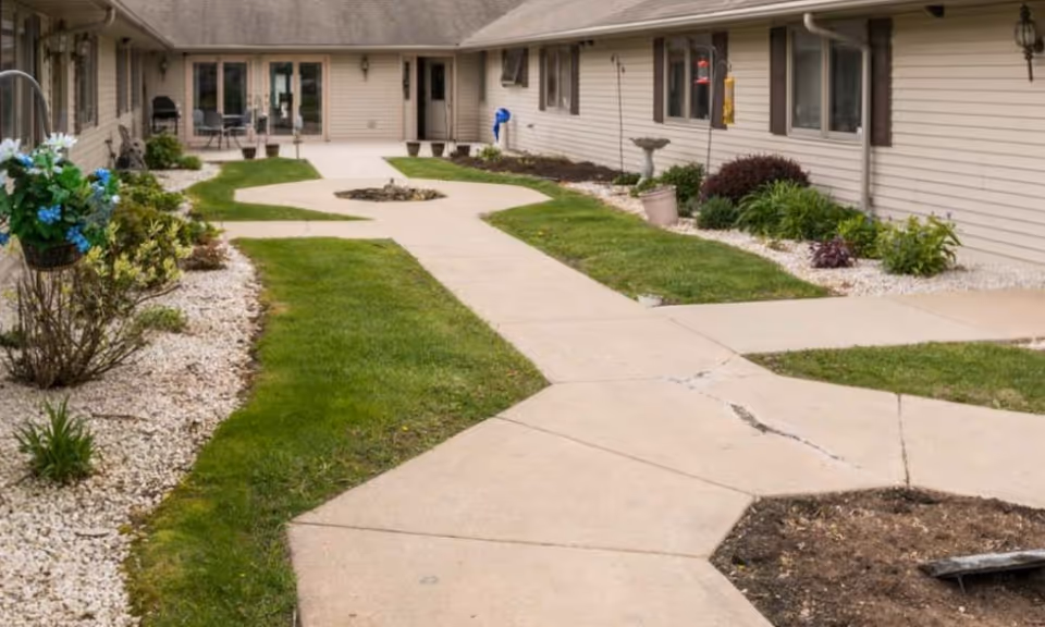 Paved walkway through a landscaped courtyard between single-story beige buildings with grass, planters, and outdoor seating.