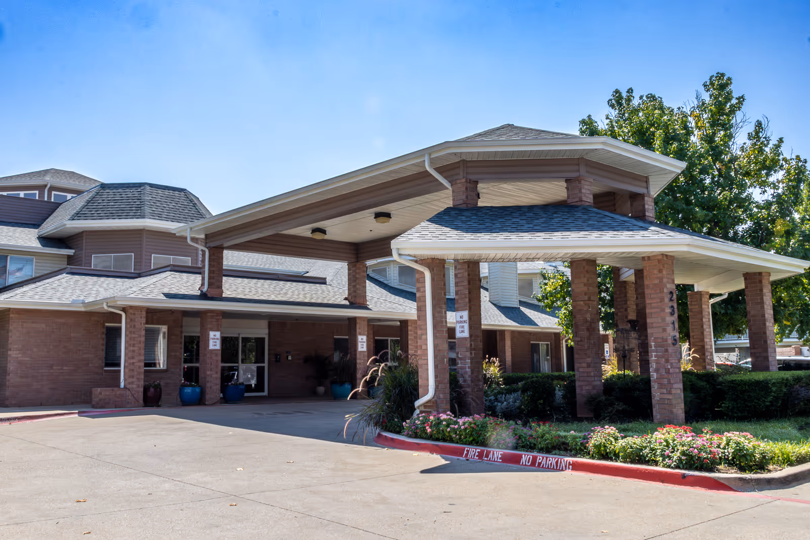 Front exterior view of a senior living facility with a covered entrance supported by brick pillars, surrounded by landscaping and a clear blue sky.