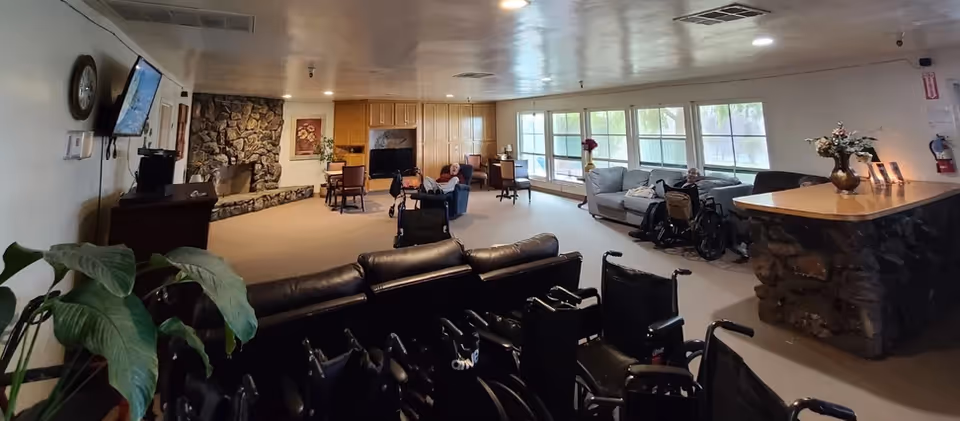 A spacious assisted living common area with several wheelchairs in the foreground, a stone fireplace on the left wall, multiple chairs and sofas arranged around the room, large windows letting in natural light, and a stone reception desk on the right side. Two elderly individuals are seated in the room, one in a wheelchair near the windows and another reclining in a chair.