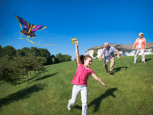 A young girl flying a colorful butterfly-shaped kite on a grassy hill with an elderly man and woman smiling and walking behind her near a residential building under a clear blue sky.