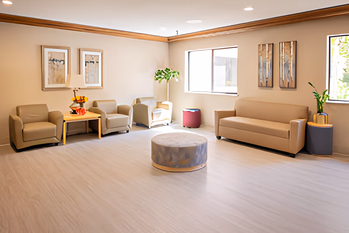A bright and spacious living room area in Porter Place Retirement Community featuring beige armchairs and a sofa arranged around a round ottoman. The room has light-colored walls with framed artwork, large windows letting in natural light, a wooden side table with a lamp and flowers, and potted plants adding greenery.