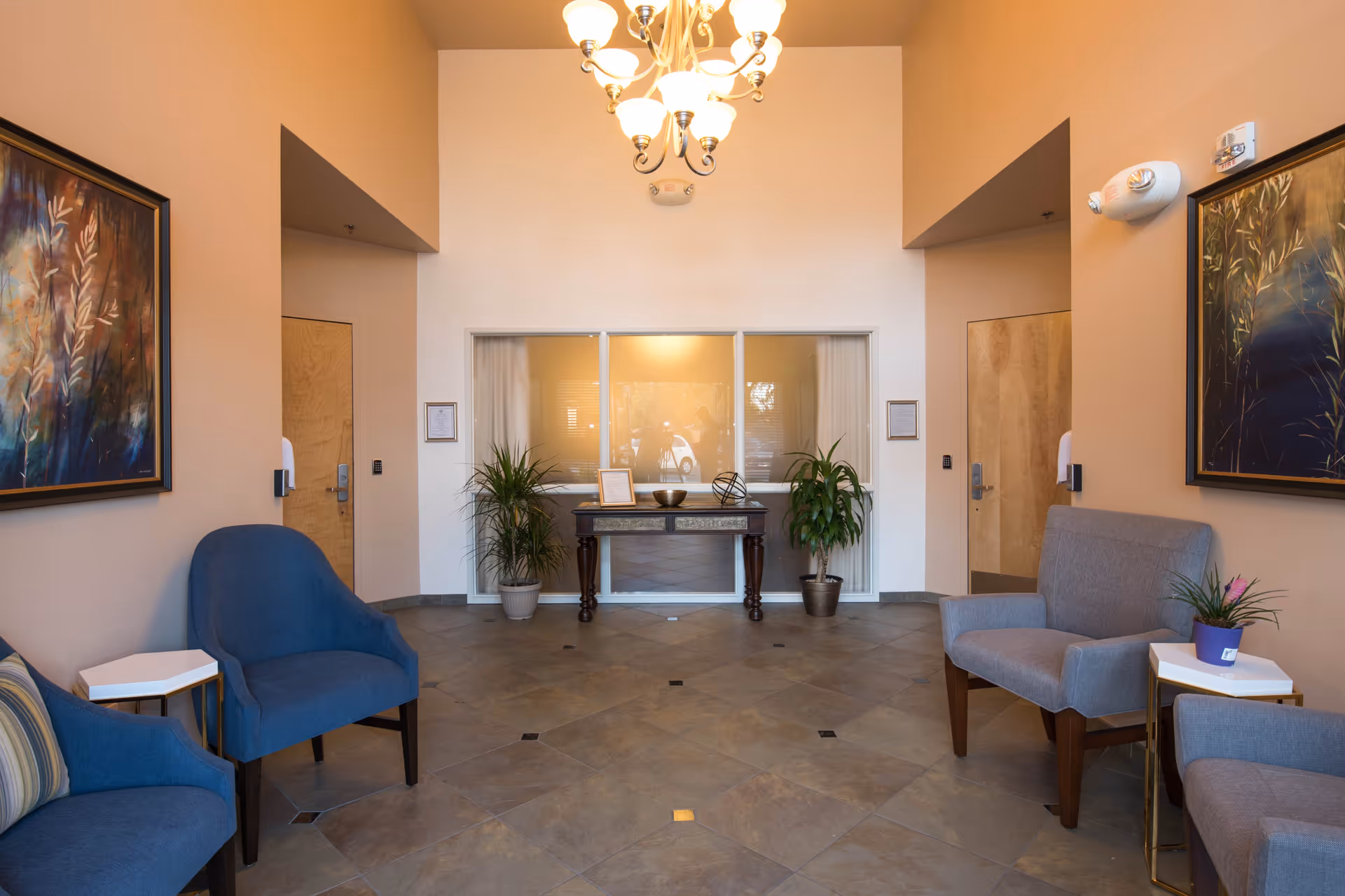 A cozy sitting area with blue and gray armchairs arranged around small white side tables, two potted plants flanking a decorative table against a wall with three windows, warm beige walls adorned with framed artwork, and a chandelier hanging from the ceiling.