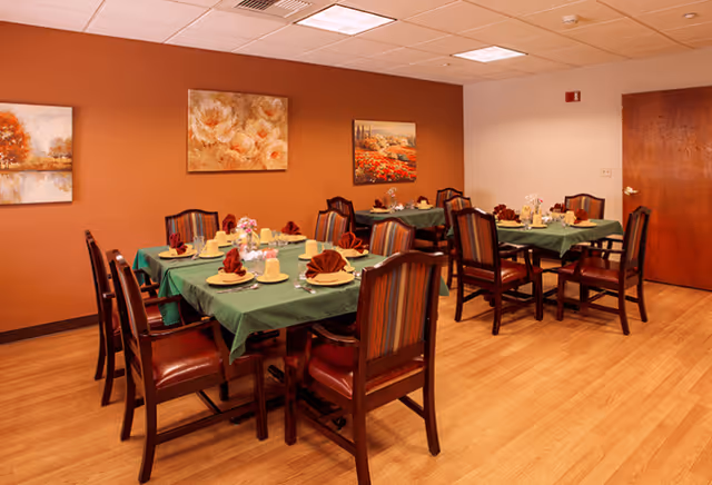 A dining room with three tables covered in green tablecloths, each set with plates, utensils, glasses, and folded red napkins. The room has wooden chairs with striped upholstery, a wooden floor, and three paintings on the orange wall. A wooden door is visible on the right side.