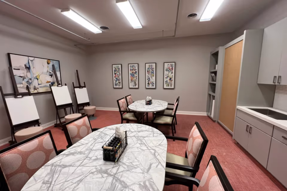 An interior activity room with marble-topped round tables, chairs, easels against the wall, and cabinets with a sink.