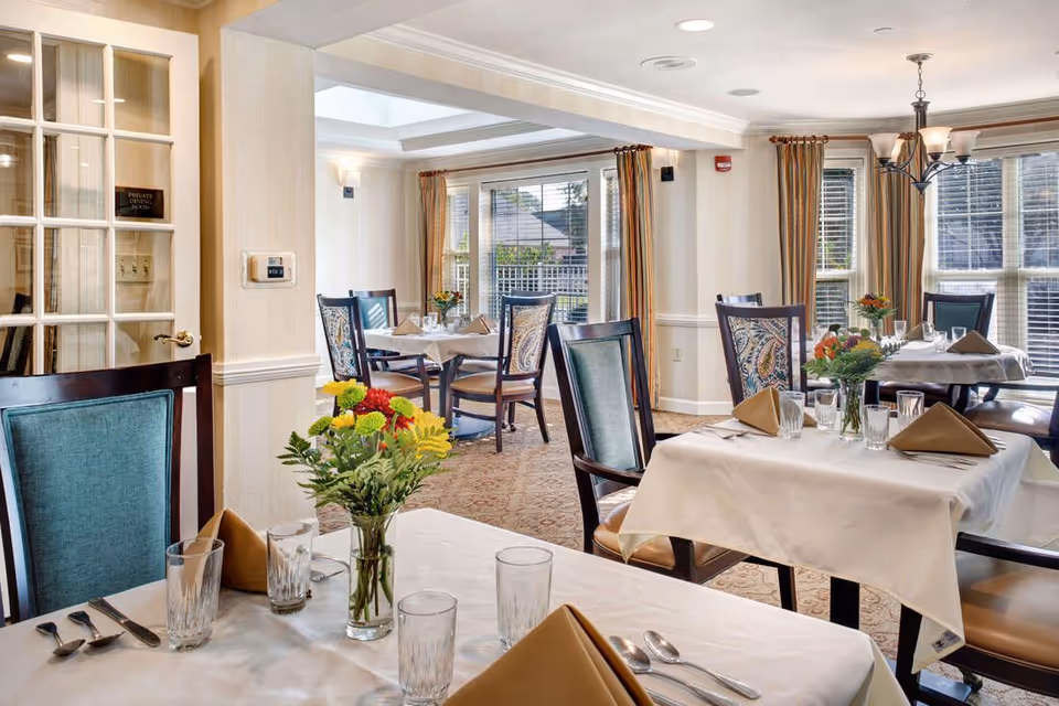 Bright dining room with tables set with white tablecloths, folded napkins, glassware, and floral centerpieces.