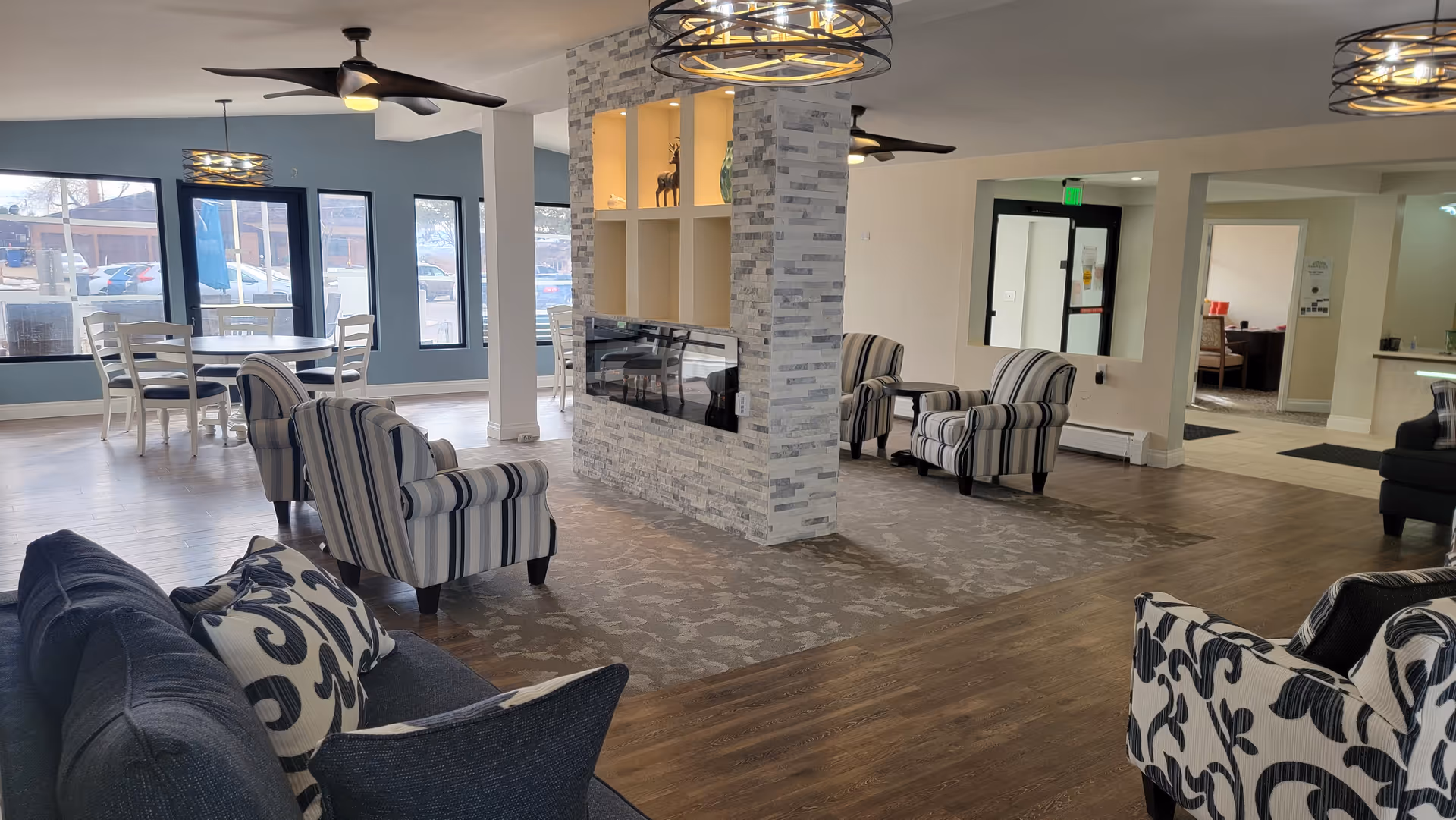 A spacious senior living facility common area with multiple seating options including striped armchairs and a dark blue couch with patterned pillows. The room features a modern stone fireplace in the center, large windows letting in natural light, ceiling fans, and a dining table with chairs near the windows. The flooring is a mix of wood and carpet, and there are open doorways leading to other rooms.