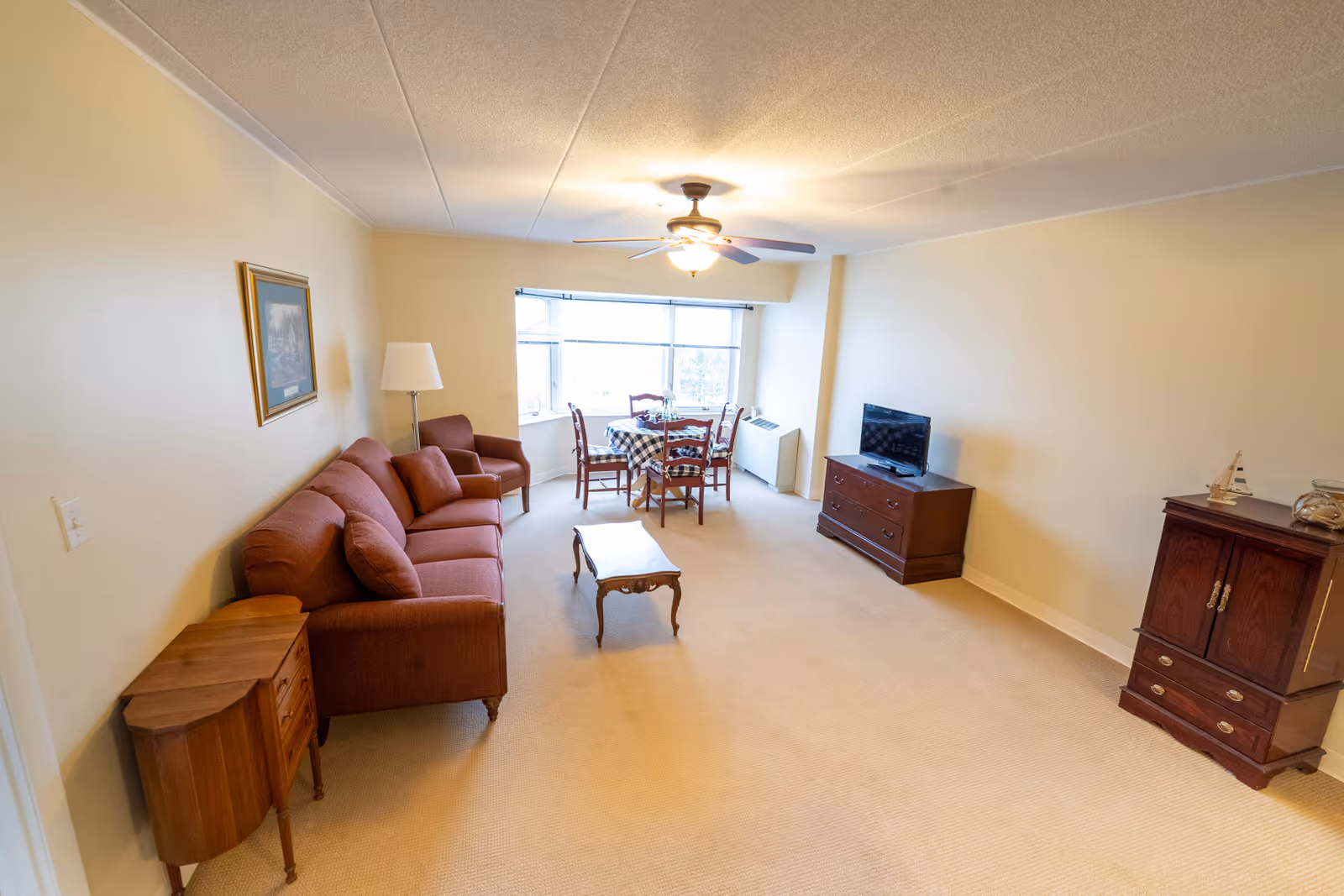 A living room with beige walls and carpet, featuring a red sofa with matching armchair, a wooden coffee table, a small wooden side table, a floor lamp, a dining table with four chairs near a large window, a flat-screen TV on a wooden dresser, and a wooden cabinet with decorative items.