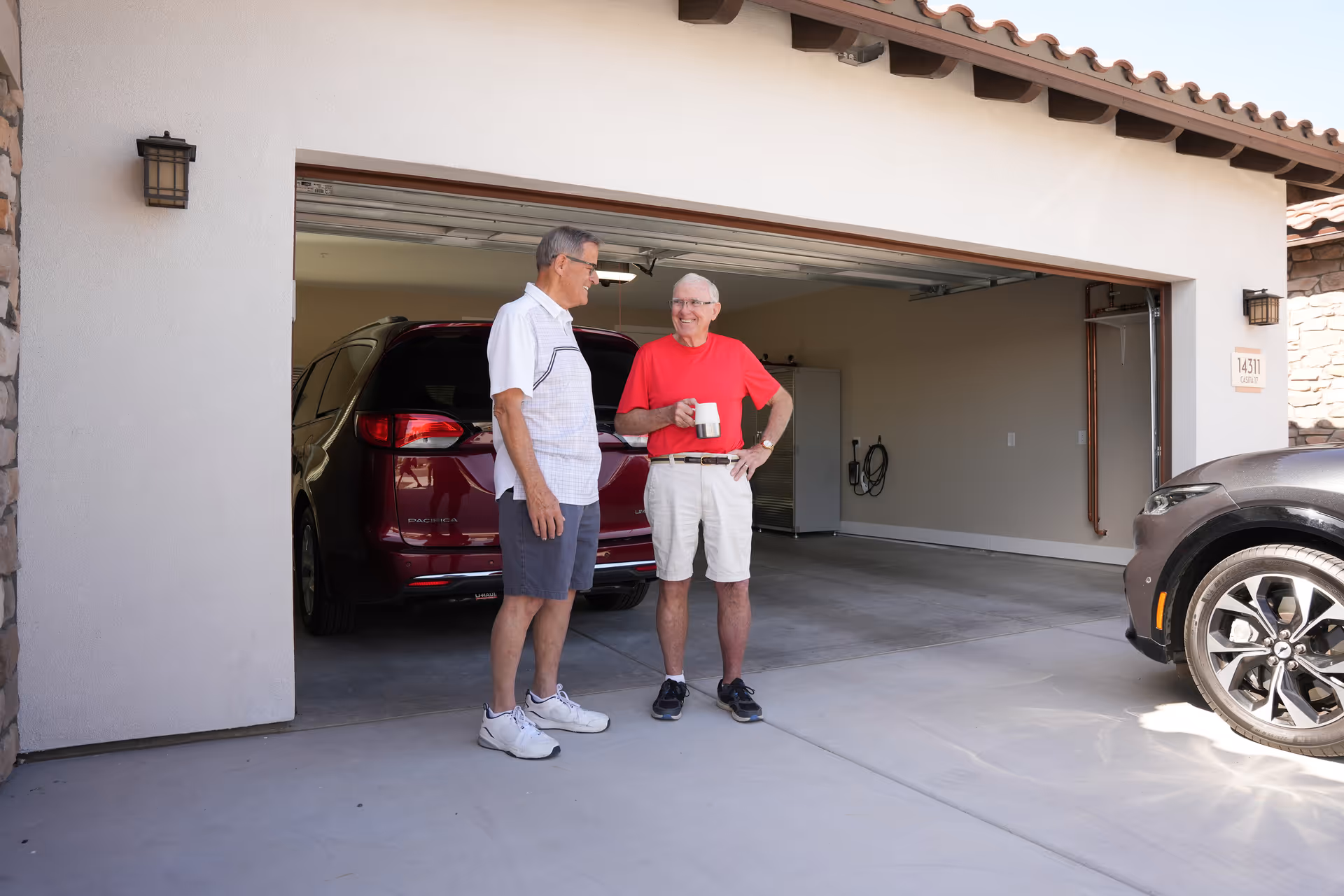 Two elderly men standing and talking in front of an open garage with a red SUV parked inside. One man is wearing a white shirt and shorts, and the other is wearing a red shirt and light-colored shorts while holding a coffee mug. A black car is partially visible on the right side of the image.
