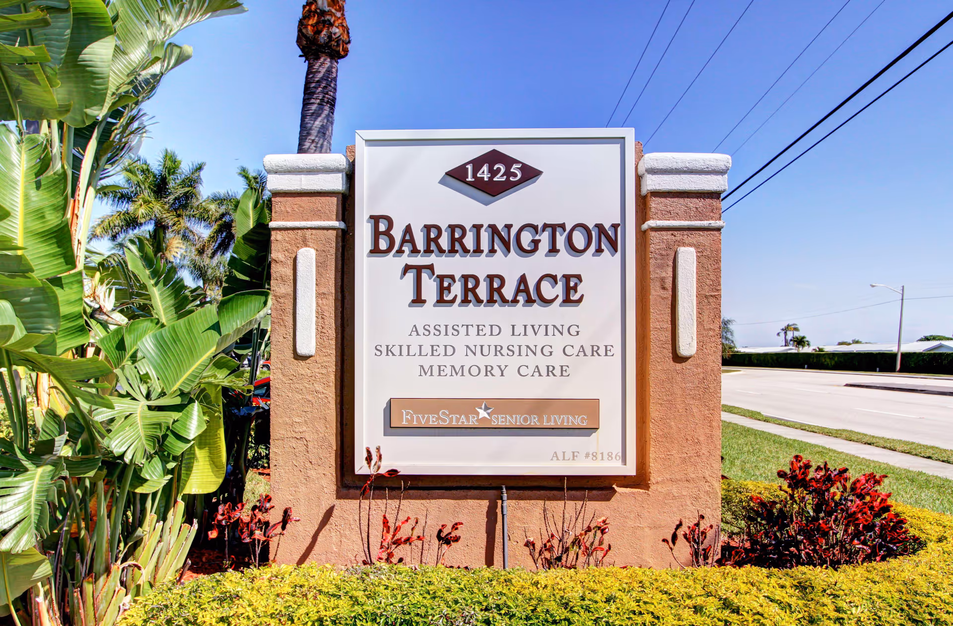 Entrance sign reading 'Barrington Terrace' with landscaping, palm trees, and a street in the background.