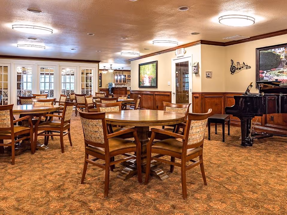 A spacious dining room with multiple round wooden tables and cushioned chairs arranged neatly. The room features warm lighting from ceiling fixtures, a patterned carpet, wood-paneled walls, a black grand piano, and framed artwork on the walls. There is a door with an exit sign above it and glass-paneled double doors leading to another room.