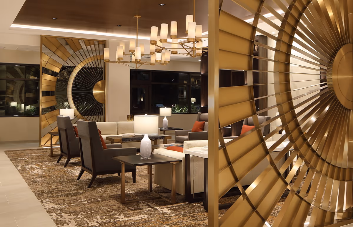 A modern and elegant living room area with beige and gray armchairs and sofas arranged around dark wood tables with white lamps. The room features a large decorative golden circular wall art piece and a matching chandelier with multiple cylindrical lampshades. The floor is covered with a patterned brown and beige rug, and large windows are visible in the background.