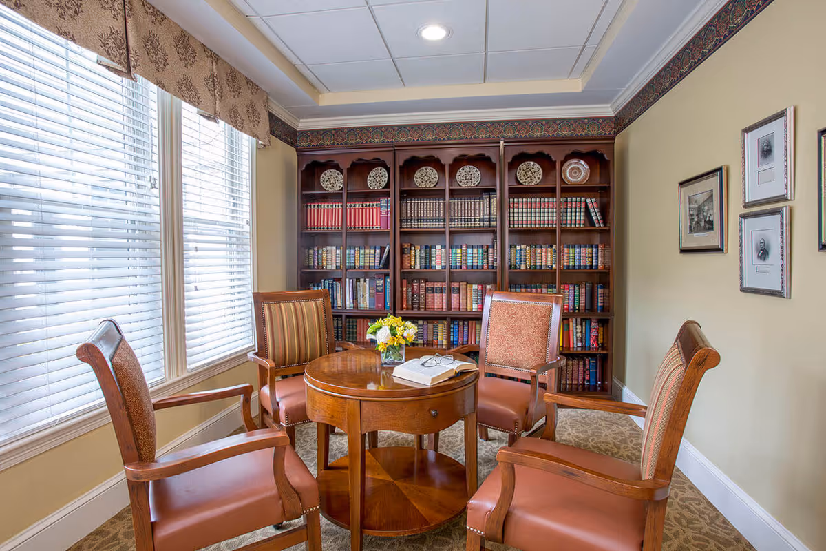A cozy reading room with four wooden armchairs upholstered in brown and patterned fabric arranged around a round wooden table. On the table, there is an open book, a pair of reading glasses, and a small vase with yellow and white flowers. Behind the table is a large wooden bookshelf filled with books and decorative plates. The room has beige walls, carpeted floor, and large windows with white blinds and patterned valances letting in natural light. Three framed black and white portraits hang on the wall to the right.