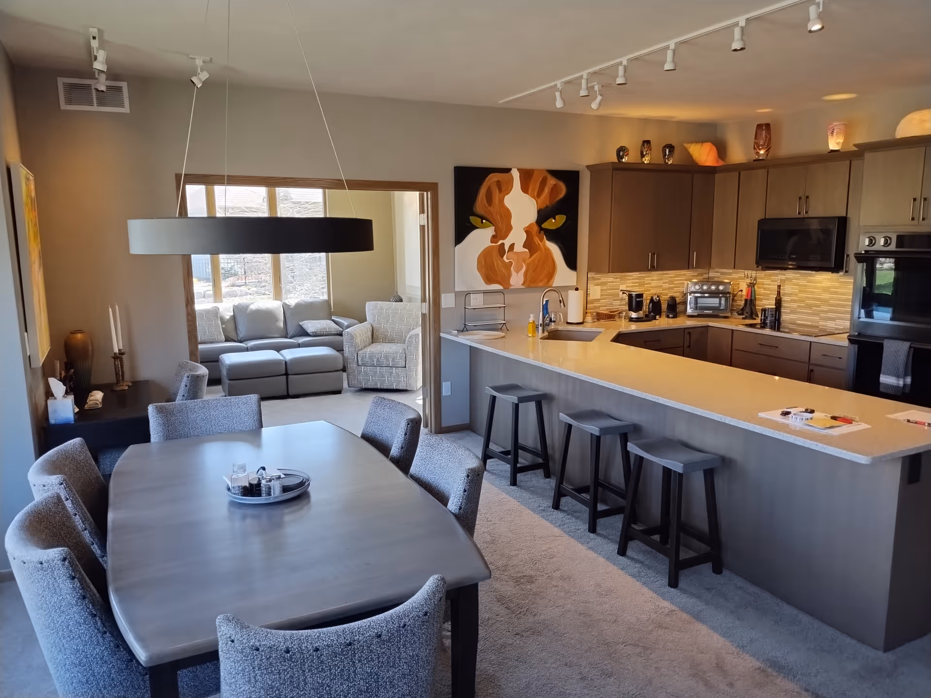 Interior view of a modern senior living facility showing a dining area with a rectangular table and six upholstered chairs, a kitchen with a long island and three bar stools, and a cozy living room with a sofa and armchair visible through an open doorway. The kitchen features wooden cabinets, a microwave, oven, and various small appliances. A large painting of a cow's face is mounted on the wall above the kitchen counter.