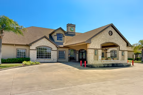 Exterior view of a single-story senior living facility building with stone and beige siding, large windows, a covered entrance, and a clear blue sky.